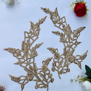 May include: Two symmetrical gold-colored embroidered floral appliques featuring intricate threadwork in a leaf and vine pattern. The designs are elongated with detailed stitching, showcasing a metallic sheen and a complex, branching botanical structure.