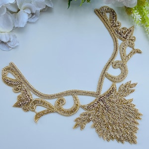 May include: Gold beaded applique with a floral design. The applique features intricate detailing with gold beads and a leaf-like pattern. The design is set against a white background, highlighting the gold colour.