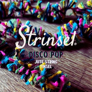 以下が含まれることがあります： カラフルなジュートストリングのクローズアップ。ピンク、ブルー、イエロー、グリーンのミックスです。「Strinsel」の文字は白いスクリプトで、その下に「DISCO POP」と「JUTE STRING TINSEL」があります。