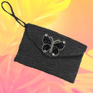 May include: A black crocheted clutch with a flap closure and a wrist strap. The flap is adorned with a butterfly design made of black beads and clear rhinestones. The background is a gradient of yellow, orange, and pink.