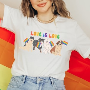 May include: A white t-shirt with the text "LOVE IS LOVE" printed on the front. Four dogs are depicted holding rainbow flags. The person wearing the shirt is holding a rainbow flag.