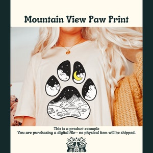 May include: A white t-shirt with a black paw print design. The paw print features a mountain range, a lake, a crescent moon, and stars. The text "Mountain View Paw Print" is at the top of the image.