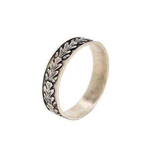 Puede incluir: Un anillo de plata con un diseño en relieve de hojas de roble y bellotas. El anillo tiene una banda con un patrón detallado de hojas y bellotas, creando una estética inspirada en la naturaleza. El anillo se muestra sobre un fondo blanco.