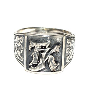 Op de afbeelding: Zilverkleurige ring met een rechthoekige voorkant met de letters "TK" in een sierlijk lettertype. De ring heeft een sierlijk ontwerp met bladeren en een getextureerd oppervlak.