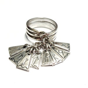 May include: A silver ring with multiple charms hanging from it. Each charm is shaped like a small shield and has a different rune engraved on it.