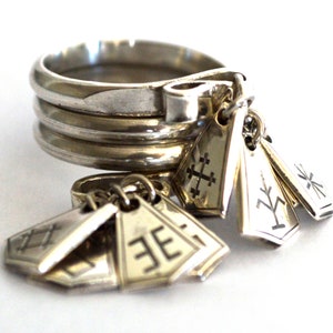 May include: A silver ring with multiple bands and several charms hanging from it. The charms are shaped like small rectangular tags with different symbols engraved on them.