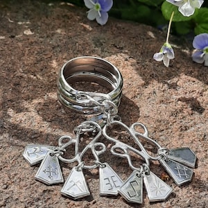 May include: A silver ring with multiple bands connected to a charm with several small, silver pendants. Each pendant has a different engraved design. The ring and charms are displayed on a textured, brown surface with small purple flowers in the background.