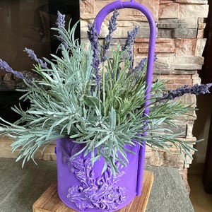 May include: A purple metal planter with a decorative floral design, holding lavender and other greenery. The planter has a curved handle and sits on a wooden base. The overall design is a decorative floral arrangement.