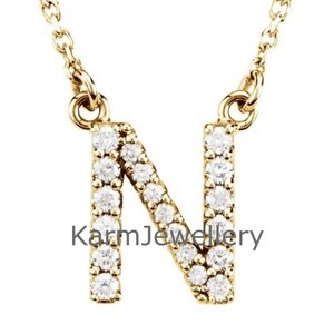 May include: A gold-coloured necklace featuring an "N" pendant. The pendant is set with small, clear gemstones. The necklace chain is delicate and gold-coloured. The text "KarmJewellery" is printed on the pendant.