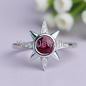 May include: A silver ring featuring a starburst design with a central, round, deep red gemstone. The starburst has pointed edges and is accented with small, clear gemstones. The ring is set against a soft purple background.