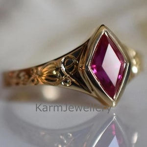May include: A gold ring featuring a diamond-shaped, ruby-red gemstone. The band is decorated with detailed floral and swirl patterns. The text "KarmJewellery" is visible on the ring.