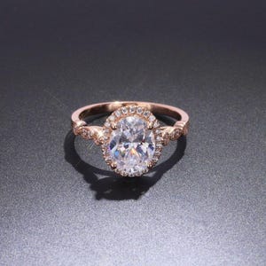 May include: A rose gold ring featuring a large oval-cut center stone surrounded by a halo of small round accent stones. The band is crafted with a delicate, scalloped metal design that extends along the sides, set with additional small round stones.