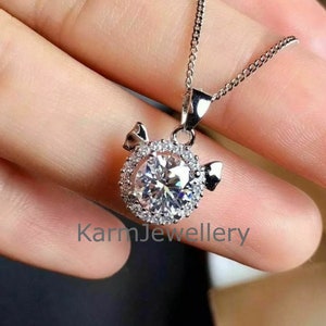 May include: A silver necklace with a round pendant featuring a large clear crystal surrounded by smaller crystals. The pendant is shaped like a pig with small wings on either side. The text "KarmJewellery" is visible at the bottom of the image.