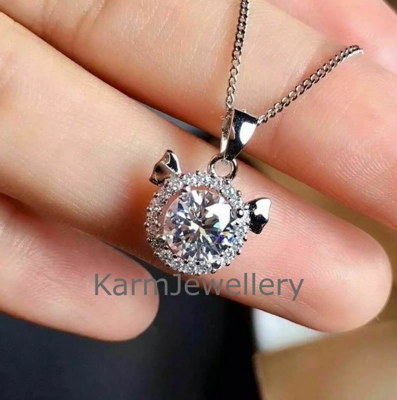 May include: A silver necklace with a round pendant featuring a large clear crystal surrounded by smaller crystals. The pendant is shaped like a pig with small wings on either side. The text "KarmJewellery" is visible at the bottom of the image.