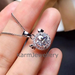 May include: A silver necklace with a pendant featuring a large, round, clear gemstone surrounded by smaller, clear gemstones. The pendant is shaped like a pig with small ears. The text "karmJewellery" is visible in the image.