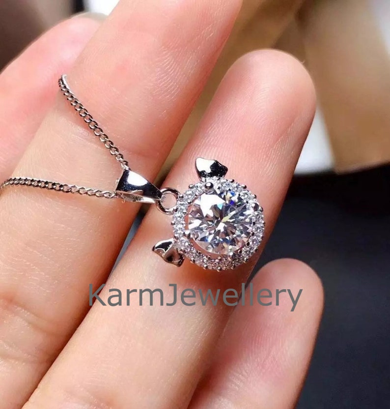 May include: A silver necklace with a pendant featuring a large, round, clear gemstone surrounded by smaller, clear gemstones. The pendant is shaped like a pig with small ears. The text "karmJewellery" is visible in the image.