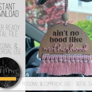 May include: A wooden car air freshener with a pink tassel. The air freshener is shaped like a half circle and has the text "ain't no hood like motherhood" written on it.