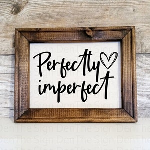 May include: A framed sign with the words "Perfectly imperfect" in black script, with a heart symbol. The sign has a white background and a dark brown wooden frame. The sign is a decorative item.