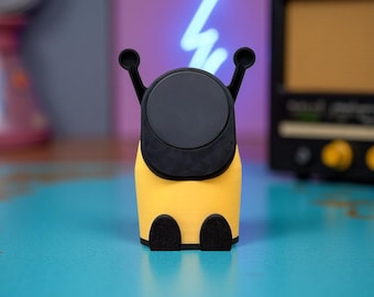 May include: A yellow and black phone accessory with a circular black top and two black antennae-like features. The accessory has two black feet and is set against a blurred background.
