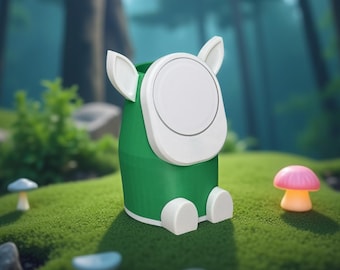 May include: A whimsical green and white container with a rounded top and ear-like protrusions, resembling a cartoon character. It sits on a bed of green moss, surrounded by miniature mushroom decorations. The background features a blurred forest scene.