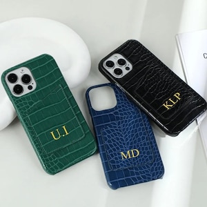 Puede incluir: Tres fundas para teléfono en verde, negro y azul, cada una con un patrón de cocodrilo texturizado. Las fundas tienen una ranura para tarjetas en la parte posterior e iniciales doradas: U.I, KLP y MD. Las fundas están sobre una superficie blanca.