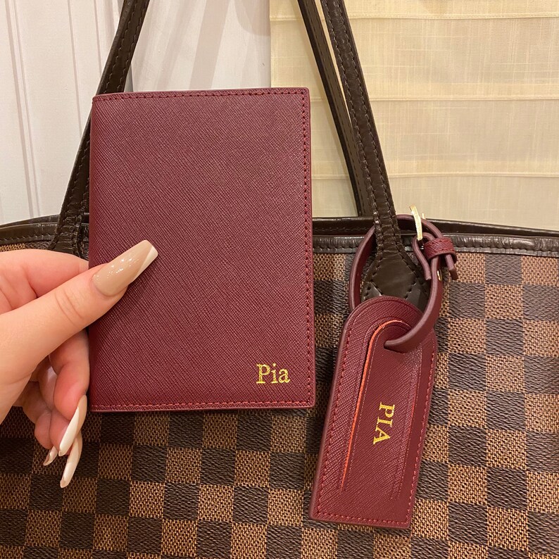 May include: A burgundy leather passport holder with the name "Pia" embossed in gold lettering. The holder is attached to a brown and tan checkered tote bag with a burgundy leather luggage tag that also says "Pia" in gold lettering.