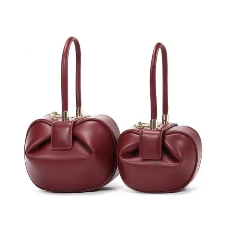 May include: Two burgundy leather handbags with a unique rounded shape and gold hardware. The handbags have top handles and a slightly puffed design.