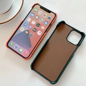 May include: Two phone cases, one orange and one green. The orange case has a cutout for the camera lens and is made of a soft, textured material. The green case has a brown leather-like interior and a green outer edge.