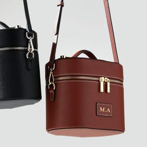 May include: A cylindrical handbag in dark brown with a textured finish, featuring a top handle, a detachable shoulder strap, gold-tone hardware, a double zipper closure, and a rectangular patch on the front with the gold embossed letters M.A.