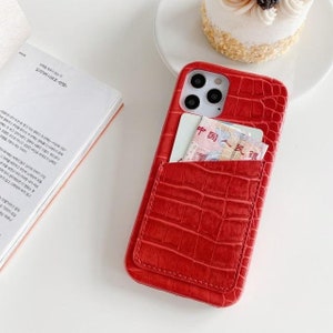 May include: Red phone case with a crocodile pattern and a pocket on the back for a card or cash. The case has a camera cutout and a black background.