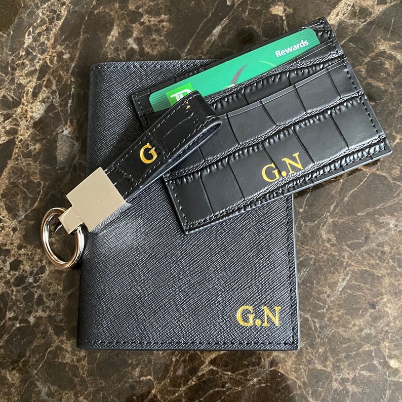 May include: Black leather wallet with a card holder and a key chain. The wallet has the initials "G.N." embossed in gold on the front. The card holder has a green credit card with the word "Rewards" printed on it.