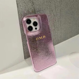 Puede incluir: Una funda de teléfono rosa con un acabado texturizado y una inscripción dorada que dice "D.M.B."