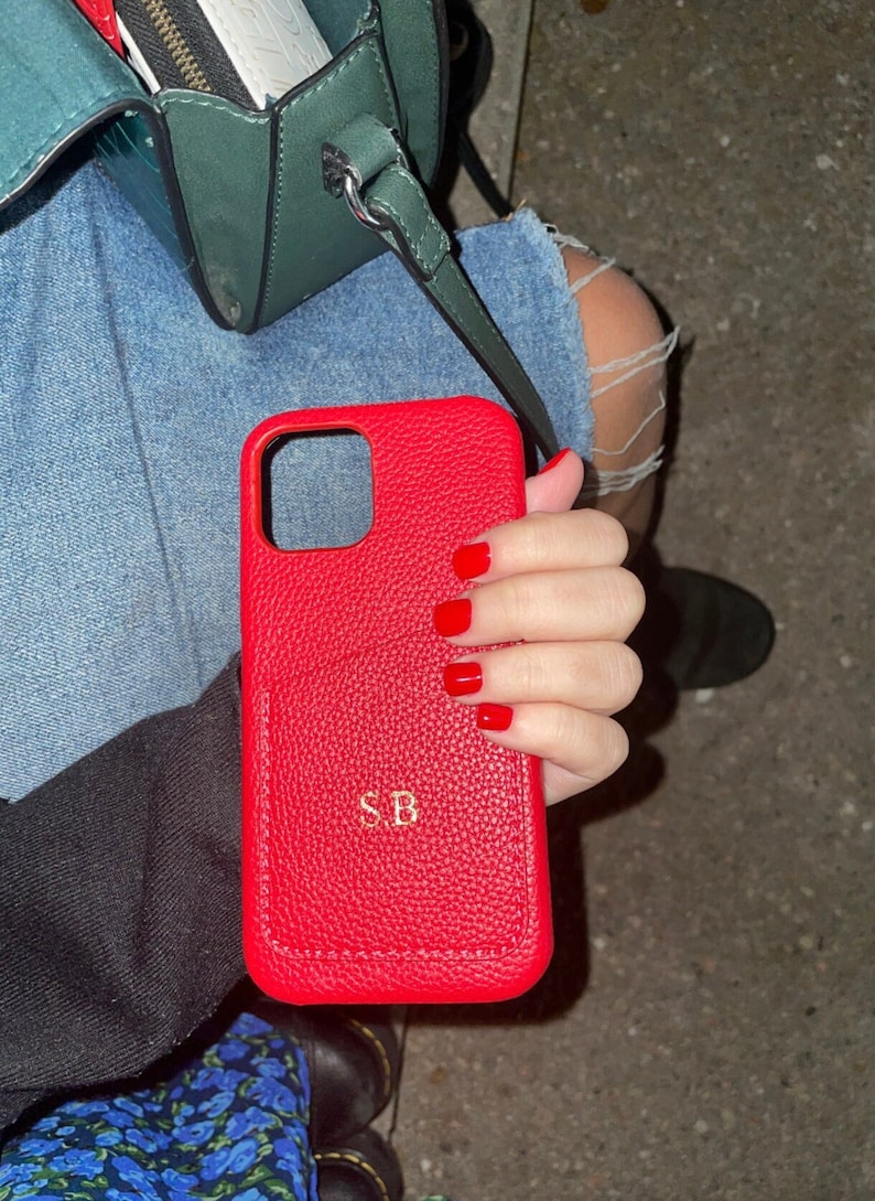 May include: Red leather phone case with the initials "SB" embossed in gold. The case has a textured surface and is held in a hand with red nail polish.