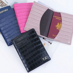 May include: Four passport holders in different colors: navy blue, pink, light brown, and black. The black passport holder has the initials "T.C." embossed in gold. The passport holders have a crocodile-embossed leather design.
