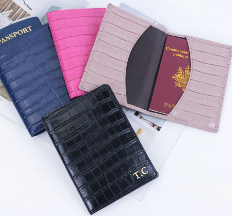 May include: Four passport holders in different colors: navy blue, pink, light brown, and black. The black passport holder has the initials "T.C." embossed in gold. The passport holders have a crocodile-embossed leather design.