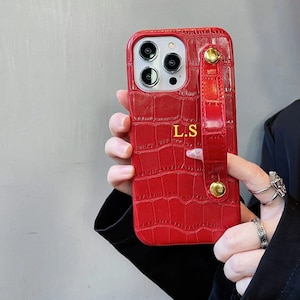 May include: A red phone case with a crocodile skin texture and a matching wrist strap. The case has gold-colored hardware and the initials "L.S" in gold. The phone case is designed to fit a smartphone.