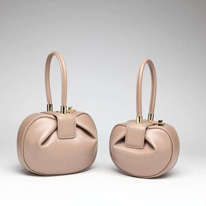 K&ouml;nnte beinhalten: Zwei beigefarbene Ledertaschen mit goldenen Beschl&auml;gen und runder Form. Die Handtaschen haben einen einzigen oberen Griff.