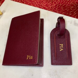 May include: Burgundy leather passport holder and luggage tag set with gold lettering. The passport holder has the name "Pia" embossed on the front. The luggage tag has the name "PIA" embossed on the front.