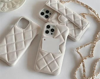 Puede incluir: Tres fundas de teléfono color blanco roto con patrón de diamantes acolchado. Una funda tiene un bolsillo con solapa, otra una ranura para tarjetas y la tercera es una funda estándar. Una cadena dorada larga está unida a una de las fundas.