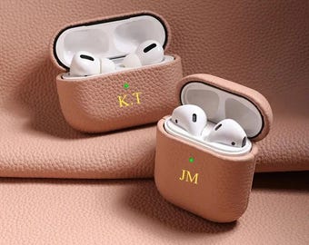 Puede incluir: Dos estuches texturizados de color rosa claro para auriculares inalámbricos, cada uno con auriculares blancos. El estuche horizontal presenta las letras doradas K.T y el vertical las letras doradas JM. Ambos tienen una luz verde.