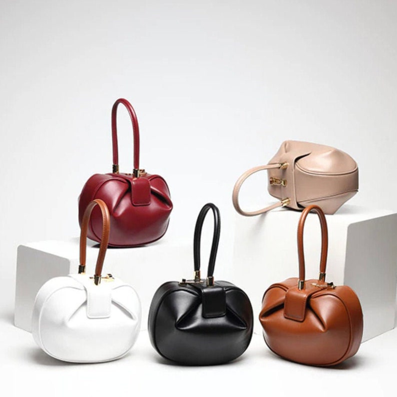 K&ouml;nnte beinhalten: F&uuml;nf verschiedene farbige, runde Ledertaschen mit einem einzigartigen, gerafften Design. Die Handtaschen haben einen oberen Griff und einen goldenen Verschluss.