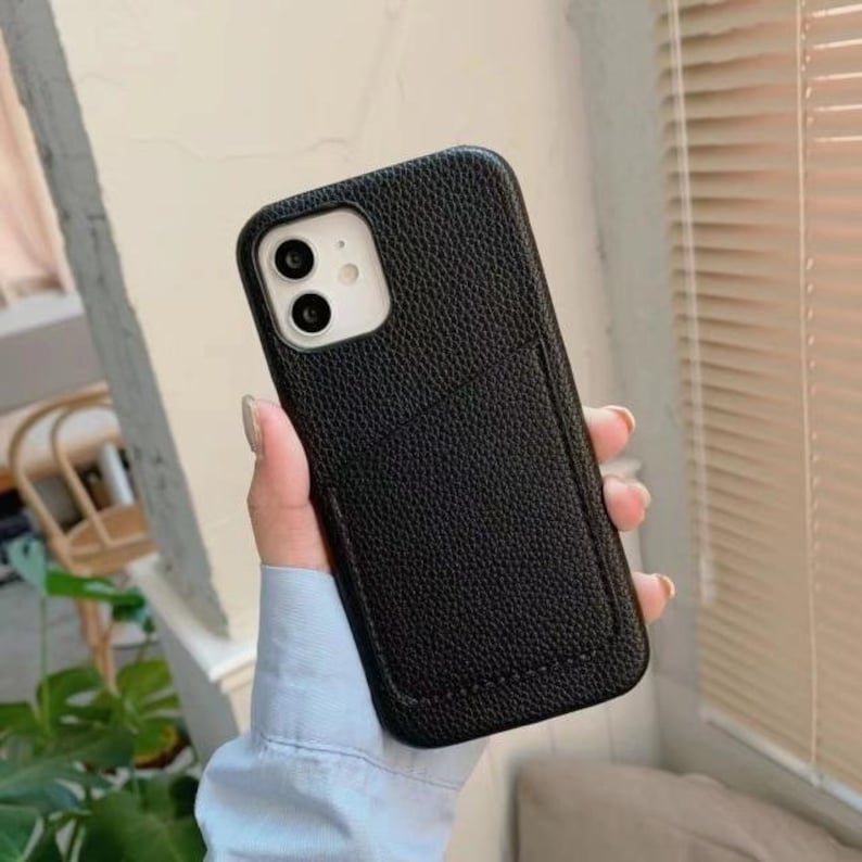 May include: Black leather phone case with a built-in card holder. The case has a textured surface and a raised lip around the camera lens.