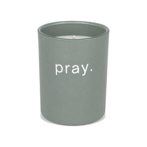 May include: A sage green cylindrical candle vessel with a matte finish. The word pray. is printed in white lowercase sans-serif letters on the front. A single white candle wick is visible in the centre of the white wax inside the container.