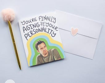 May include: A greeting card with a colourful design and the text "You're finally aging into your personality". The card features a photo of a man with a surprised expression.