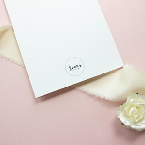 May include: A white rectangular card featuring a circular logo with the text Kacey's Creations in black script. The card is surrounded by cream-colored frayed ribbon and two small artificial flowers, one pink and one white, on a pink surface.