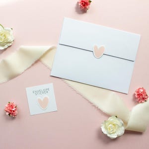 May include: A white envelope sealed with a light pink heart sticker, accompanied by a square card labeled ENVELOPE STICKER with a matching heart sticker, surrounded by cream-colored frayed ribbon and small white and pink artificial flowers.