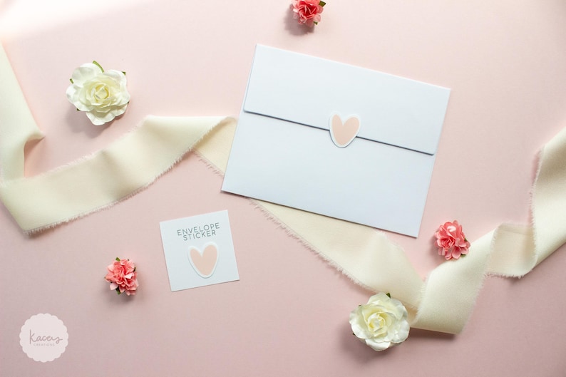 May include: A white envelope sealed with a light pink heart sticker, accompanied by a square card labeled ENVELOPE STICKER with a matching heart sticker, surrounded by cream-colored frayed ribbon and small white and pink artificial flowers.