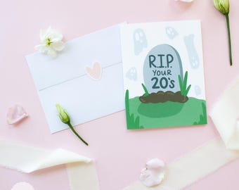 May include: A pink and white greeting card featuring a cartoon illustration of a tombstone with the text "RIP...your 20's". The card is surrounded by white flowers and a white ribbon.