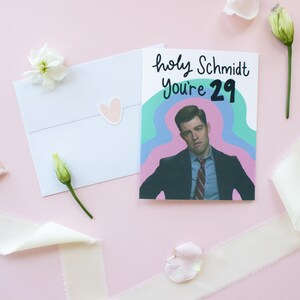 May include: A white greeting card featuring a graphic illustration of a man in a suit and tie with the text "holy Schmidt You're 29" in black lettering against a wavy teal and purple background, accompanied by a white envelope with a heart sticker.