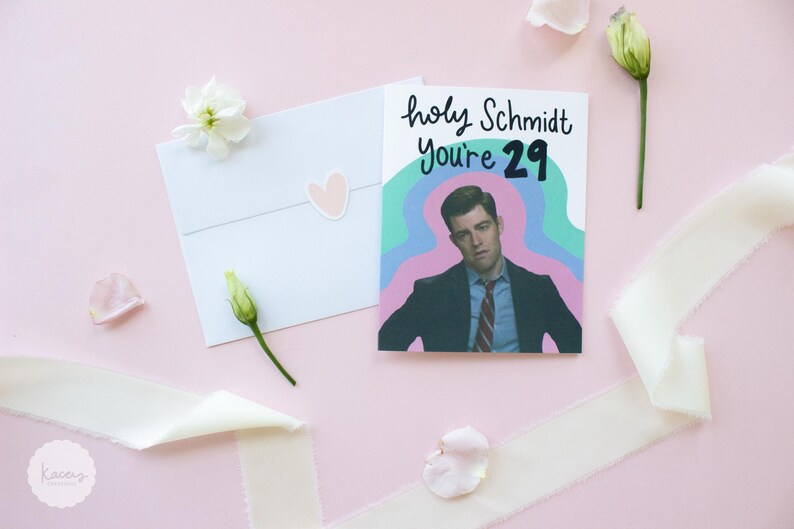 May include: A white greeting card featuring a graphic illustration of a man in a suit and tie with the text "holy Schmidt You're 29" in black lettering against a wavy teal and purple background, accompanied by a white envelope with a heart sticker.